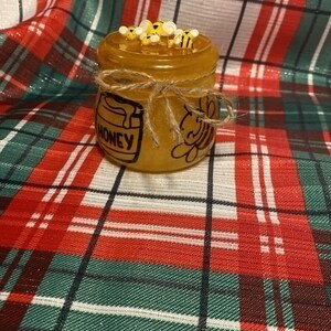 Puede incluir: Un tarro de vidrio lleno de miel, decorado con una abeja y la palabra "HONEY". El tarro está sobre una tela de cuadros rojos y verdes.