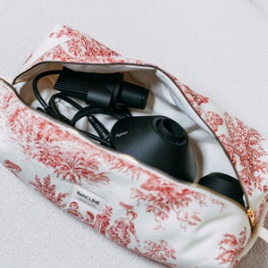 Può includere: Una borsa da toilette con cerniera in tessuto bianco con motivo toile de jouy rosso e una piccola etichetta rettangolare con la scritta BANCLINE. All'interno della borsa aperta ci sono accessori neri per capelli e un asciugacapelli Dyson.