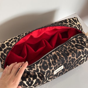 May include: A rectangular zippered pouch featuring a black and brown leopard print pattern on the exterior. The interior is lined with solid red fabric. A small white rectangular label with the text BANCLINE is sewn onto the front exterior.