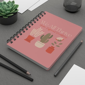 May include: A spiral-bound notebook with a dusty rose cover featuring the text We All Thrive Under Different Conditions and illustrations of three potted plants including a leafy plant, a cactus, and a flower in terracotta and cream pots.