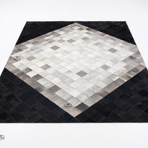 May include: A square rug featuring a central diamond pattern composed of small, light grey and white square tiles, surrounded by a border of solid black square tiles. The rug has a repeating circular logo stamp visible on the surface.