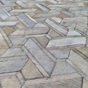 May include: A large, textured rug with a geometric pattern. The rug is made of various rectangular and trapezoidal pieces of hide, stitched together. The color palette is neutral, with shades of gray, beige, and cream.