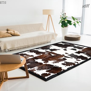 Puede incluir: Una alfombra rectangular de piel de vaca con borde negro. La alfombra presenta un patrón de manchas marrones y negras sobre un fondo blanco. Las palabras "MBM CARPETS" están impresas en la alfombra.
