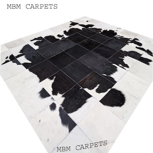 Könnte beinhalten: Ein Patchwork-Kuhfellteppich mit einem auffälligen Schwarz-Weiß-Muster. Der Teppich besteht aus quadratischen und rechteckigen Teilen, die ein geometrisches Design bilden. Der Text "MBM CARPETS" ist oben und unten im Bild schwarz gedruckt.