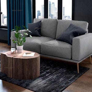 Puede incluir: Un salón moderno con un sofá gris y dos cojines azul marino. Una mesa de centro de tronco de madera se encuentra sobre una alfombra oscura, con una planta en maceta y un tarro de cristal. La habitación tiene grandes ventanas y cortinas azules.