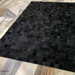 May include: A rectangular black rug with a geometric pattern of squares. The rug is made of a soft, textured material and is displayed on a light-colored stone floor. The rug's dark color provides a striking contrast to the lighter background.