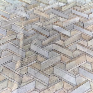 May include: A close-up view of a rug with a geometric pattern. The rug is made of rectangular pieces of varying shades of beige, gray, and white, stitched together. The pattern creates a textured, three-dimensional effect.