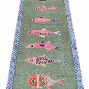 Op de afbeelding: Een rechthoekig groen tapijt met een blauwe rand en franjes, met kleurrijke visontwerpen. De vissen variëren in patroon en kleur, waaronder roze, zwart en oranje, wat een speels en decoratief accent creëert.
