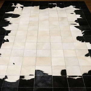 May include: A rectangular area rug constructed from individual square patches of cowhide. The center features a large block of cream and white squares, while the border consists of solid black and mottled black and white cowhide squares.
