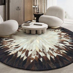 Op de afbeelding: Een groot rond vloerkleed met een geometrisch sterpatroon bestaande uit aan elkaar genaaide koeienhuidstukken in tinten wit, beige, bruin en zwart, uitstralend vanuit het midden naar de donkere buitenrand van het kleed.