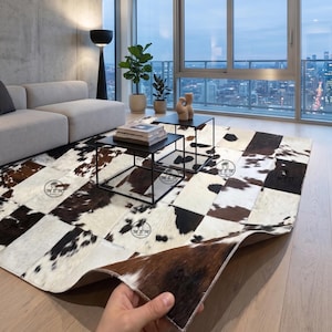 Peut inclure: Un tapis rectangulaire composé d'un assemblage de pièces de peau de vache dans des tons marron, noir et blanc. Le tapis est posé sur un parquet clair, avec un coin soulevé pour montrer le dessous et les bords cousus de l'ouvrage.