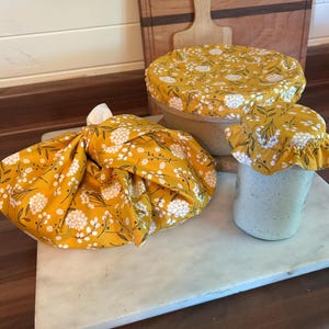 May include: Three fabric bowl covers featuring a mustard yellow background with a white and black floral pattern. One cover is tied over a bowl, one is stretched over a larger bowl, and one is fitted with a ruffled edge over a glass jar.