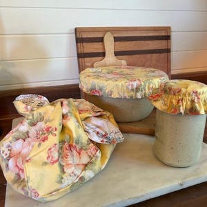 May include: Three kitchen storage items featuring a yellow fabric with a pink and white floral pattern. One item is a fabric-covered bowl, another is a fabric-covered jar, and the third is a fabric bundle tied with a knot, all displaying matching floral prints.