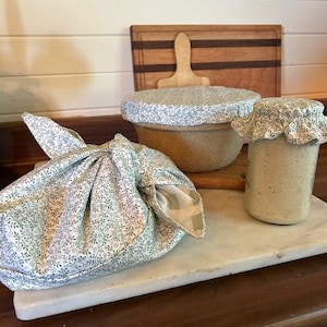 May include: A set of three fabric bowl and jar covers featuring a green and white botanical vine pattern. One cover is tied in a knot over a bowl, another is stretched over a bowl, and the third is secured with an elastic band over a glass jar.
