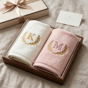 Op de afbeelding: Een geschenkset met twee opgerolde handdoeken, één witte en één roze, in een houten dienblad. Elke handdoek is geborduurd met een gouden lauwerkrans en een enkele initiaal, "K" op de witte handdoek en "M" op de roze handdoek. Een geschenkdoos met een lint staat op de achtergrond.