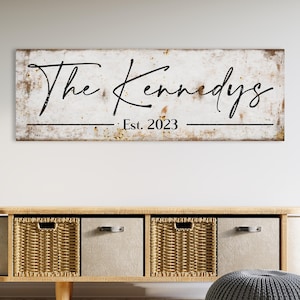 May include: A rectangular wooden sign with the text "The Kennedys" in a cursive font, with "Est. 2023" below. The sign has a distressed, vintage look with a white background and black lettering. The sign is mounted above a wooden cabinet with woven baskets and fabric drawers.