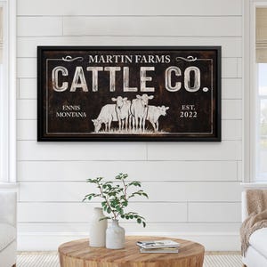 Może przedstawiać: Oprawiony znak z napisem "MARTIN FARMS CATTLE CO." w białych literach. Znak przedstawia ilustrację kilku kr&oacute;w oraz napis "ENNIS MONTANA EST. 2022" w kolorze białym. Znak ma ciemnobrązowe tło i czarną ramę.