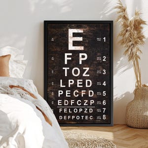 May include: A framed eye chart with a dark brown background and white letters. The chart includes the letters E, F, P, T, O, Z, L, and D, arranged in descending sizes. The chart is in a black frame.