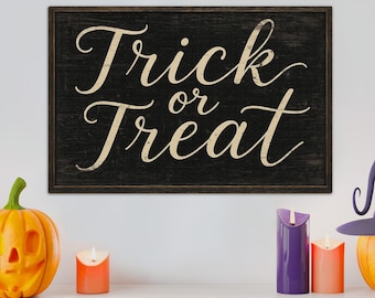 May include: A rectangular wooden sign with the words "Trick or Treat" in a cream-colored script font. The sign is set against a black background. The scene includes a carved pumpkin, candles, and a witch's hat.