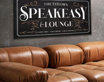 May include: A rectangular black sign with white text that reads "THE TAYLOR'S SPEAKEASY & LOUNGE." The sign also includes the phrases "GOOD TIMES • COLD DRINKS • GREAT FRIENDS." Below the sign is a brown leather couch.