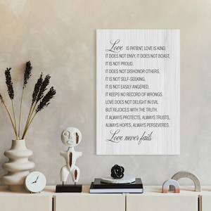 May include: A white wooden sign with the text "Love" in elegant script, listing qualities of love. The sign is part of a decorative display on a light-colored cabinet, alongside a vase with dried plants and a small sculpture.