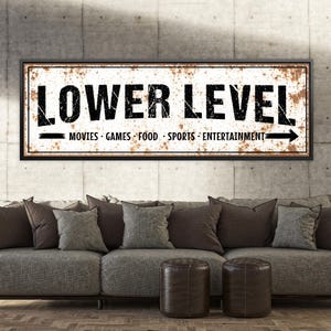 May include: A rectangular sign with the words "LOWER LEVEL" in bold, black letters. Below, it lists "MOVIES GAMES FOOD SPORTS ENTERTAINMENT" with an arrow. The sign has a distressed, rusty appearance and is framed in black.