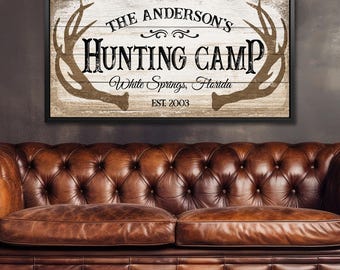 Puede incluir: Un letrero de pared rectangular con un fondo de tablones de madera blanca desgastada y un marco negro. El letrero presenta ilustraciones de astas de ciervo marrones en los lados y texto negro que dice THE ANDERSON'S HUNTING CAMP, White Springs, Florida, EST. 2003.