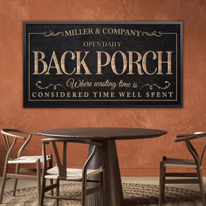 May include: A framed sign with the text "BACK PORCH" in large, embossed letters. The sign also includes the text "MILLER & COMPANY" and "Where wasting time is considered time well spent." The sign is displayed above a wooden table and chairs.