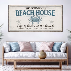 May include: A rectangular wooden sign with the text "THE JOHNSON'S BEACH HOUSE" in blue, featuring a crab and starfish. The sign also reads "Life is better at the beach, Galveston, Texas, Established 2005."
