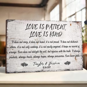 May include: A rectangular wooden sign with the text "LOVE IS PATIENT, LOVE IS KIND" in black script. The sign also includes a quote about love, names "Taylor & Jessica", and the date 12.04.2017. The sign has a distressed, white-washed finish.
