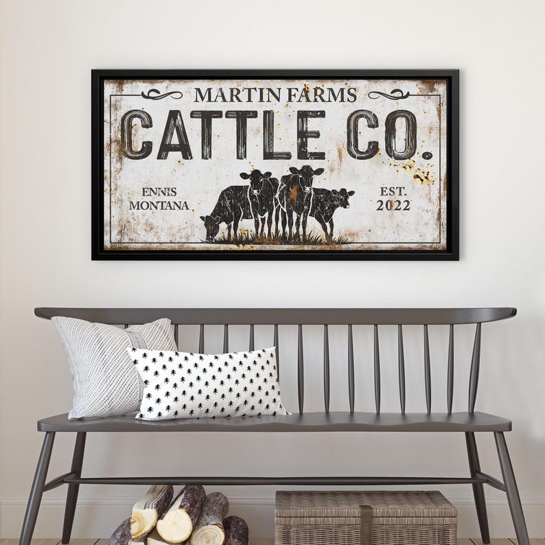 Może przedstawiać: Oprawiony znak z napisem "MARTIN FARMS CATTLE CO." i słowami "Ennis Montana Est. 2022." Znak przedstawia grafikę pięciu czarnych kr&oacute;w. Znak wisi nad szarą ławką z poduszkami.
