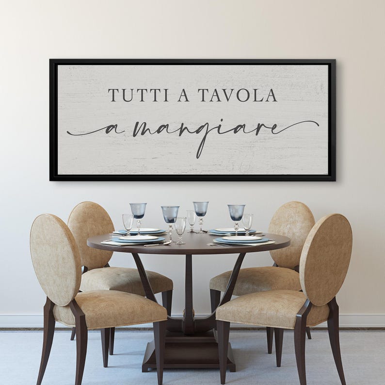 Puede incluir: Una escena de comedor con una mesa redonda de madera preparada para una comida, rodeada de seis sillas tapizadas de color beige. Encima de la mesa cuelga un letrero enmarcado con la frase italiana "TUTTI A TAVOLA a mangiare" en escritura negra.