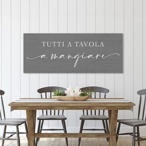 Puede incluir: Un letrero de madera gris con texto blanco "TUTTI A TAVOLA a mangiare" cuelga sobre una mesa de comedor de madera. La mesa est&aacute; puesta con un camino de mesa, velas y plantas. Cuatro sillas grises rodean la mesa.