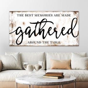 May include: A rectangular wooden sign with the text "THE BEST MEMORIES ARE MADE gathered AROUND THE TABLE" in black script. The sign has a distressed white background with brown edges, displayed above a white sofa.