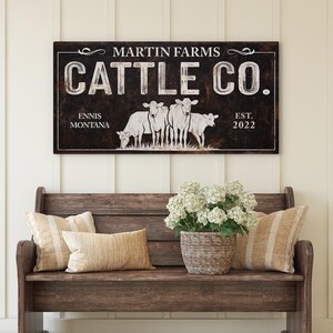 Może przedstawiać: Prostokątny drewniany znak z napisem "MARTIN FARMS CATTLE CO." i lokalizacją "ENNIS MONTANA EST. 2022." Znak przedstawia grafikę czterech kr&oacute;w. Znak wisi nad drewnianą ławką z poduszkami i koszem białych kwiat&oacute;w.