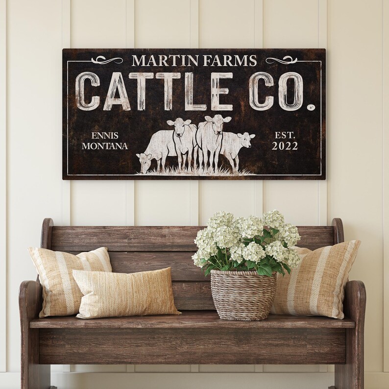 Może przedstawiać: Prostokątny drewniany znak z napisem "MARTIN FARMS CATTLE CO." i lokalizacją "ENNIS MONTANA EST. 2022." Znak przedstawia grafikę czterech kr&oacute;w. Znak wisi nad drewnianą ławką z poduszkami i koszem białych kwiat&oacute;w.