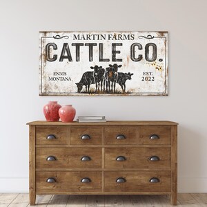 Może przedstawiać: Rustykalny drewniany szyld z napisem "MARTIN FARMS CATTLE CO." i lokalizacją "ENNIS MONTANA EST. 2022." Szyld przedstawia czarno-białą ilustrację kr&oacute;w. Jest umieszczony nad drewnianą komodą z szufladami.