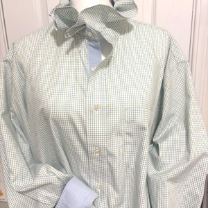 May include: A light green and white checkered shirt with a ruffled collar and light blue cuffs. The shirt has a button-down front and a classic, tailored design. The shirt is displayed on a mannequin.