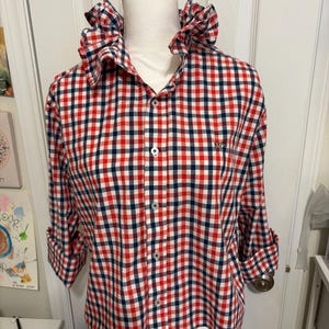 May include: A red, white, and navy blue gingham shirt with a ruffled collar and button-down front. The long sleeves are rolled up. The shirt is displayed on a white mannequin.