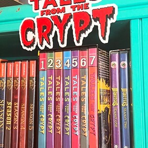 May include: A selection of DVDs, including "Tales from the Crypt" and "Monsters", displayed on a shelf. The spines are brightly coloured, with the title "Tales from the Crypt" in red and white. A sign with the show's name is above.