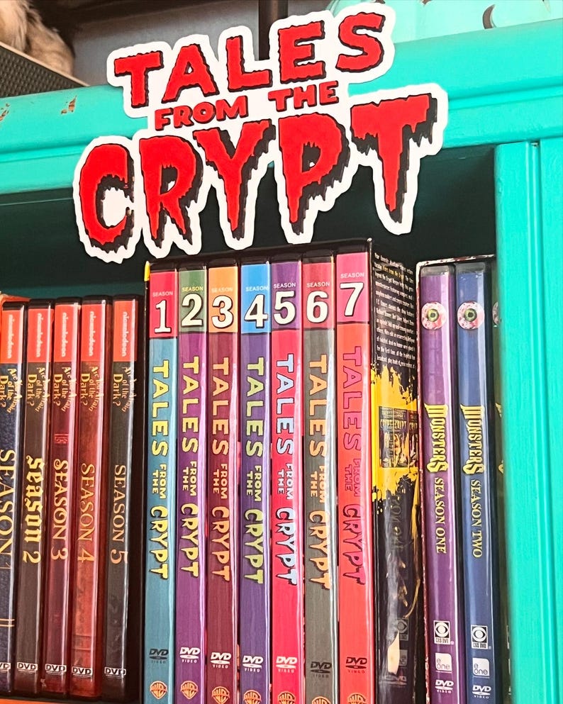 May include: A selection of DVDs, including "Tales from the Crypt" and "Monsters", displayed on a shelf. The spines are brightly coloured, with the title "Tales from the Crypt" in red and white. A sign with the show's name is above.