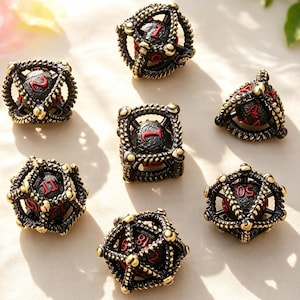 May include: Seven antique-style dice with a black and gold finish. Each die features red numbering and is encased in a decorative metal frame. The dice are arranged on a light-colored surface, suggesting a tabletop game.
