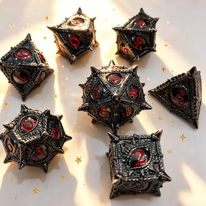 Könnte beinhalten: Eine Sammlung von sieben metallischen Würfeln mit dunkler Gunmetal-Oberfläche und roten Zahlen. Jeder Würfel hat ein einzigartiges spitzes Design, das eine gotische oder Fantasy-Ästhetik verleiht. Die Würfel sind auf einer hellen Oberfläche mit verstreuten goldenen sternförmigen Konfetti angeordnet.