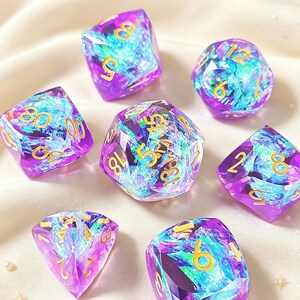 Peut inclure: Un ensemble de sept dés polyédriques composés d'une résine violette translucide incrustée de flocons de feuille irisés bleus et argentés. Chaque dé présente des bords nets et des chiffres gravés de couleur dorée pour une lecture aisée.