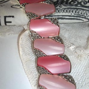 Può includere: Un braccialetto vintage con una serie di maglie rosa ovali. Ogni maglia è di una diversa tonalità di rosa, dal chiaro al rosa intenso, ed è incorniciata da un'ornata filigrana argentata. Il braccialetto è assicurato da una chiusura.