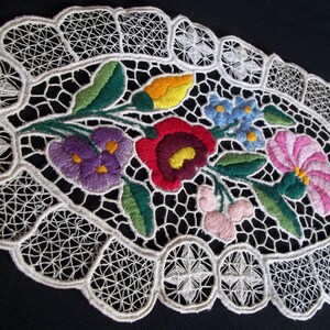 Può includere: Tovaglietta ovale in pizzo bianco con fiori ricamati colorati. Il design floreale comprende fiori viola, rossi, rosa, gialli e blu con foglie verdi. La tovaglietta ha un bordo smerlato ed è posta su uno sfondo nero.