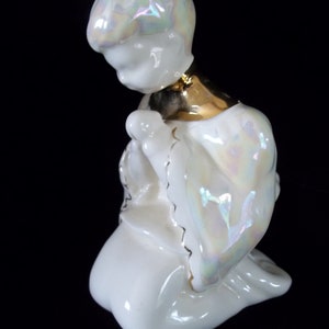 May include: A white porcelain figurine of a kneeling angel with iridescent glaze and gold accents. The angel has its head bowed and its hands clasped in prayer.