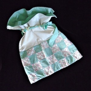 Puede incluir: Una bolsa de tela blanca con cordón de ajuste, un patrón de cuadros verdes y detalles florales. La bolsa tiene un volante de tela verde alrededor de la parte superior y una cinta verde atada alrededor de la abertura.