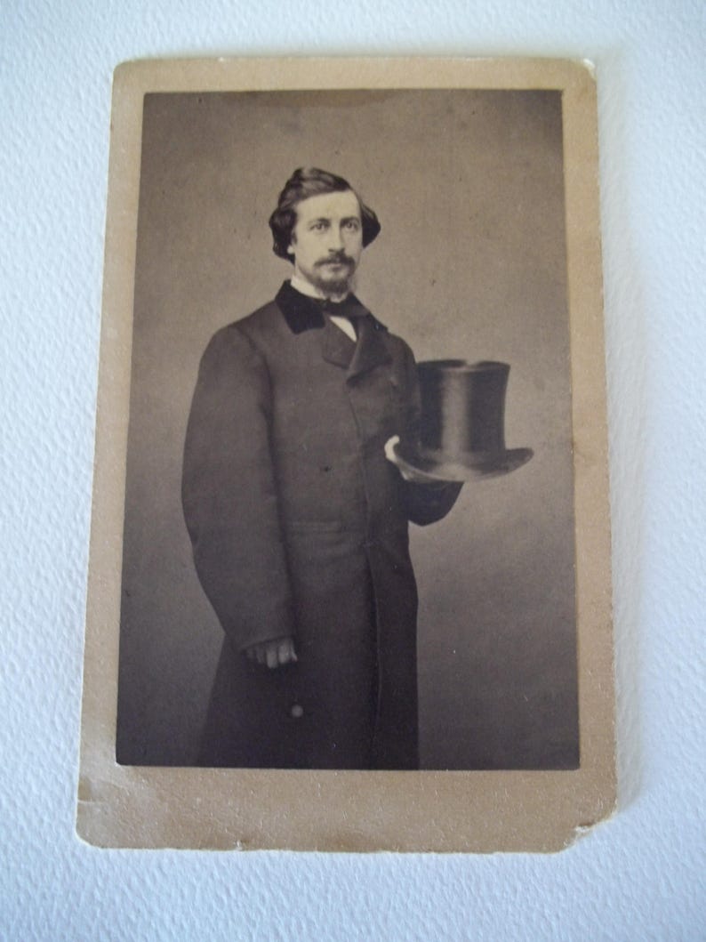 May include: Vintage black and white photograph of a man in a dark coat and top hat. The man has a mustache and is holding the hat in his right hand. The photograph has a sepia tone and is mounted on a cardboard backing.