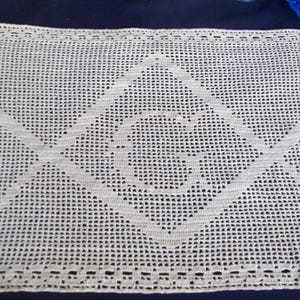 May include: Cream-colored, rectangular crocheted doily featuring a central geometric design. The design includes a diamond shape with an inner symbol, and diagonal lines extending from the diamond. The edges have a decorative border.